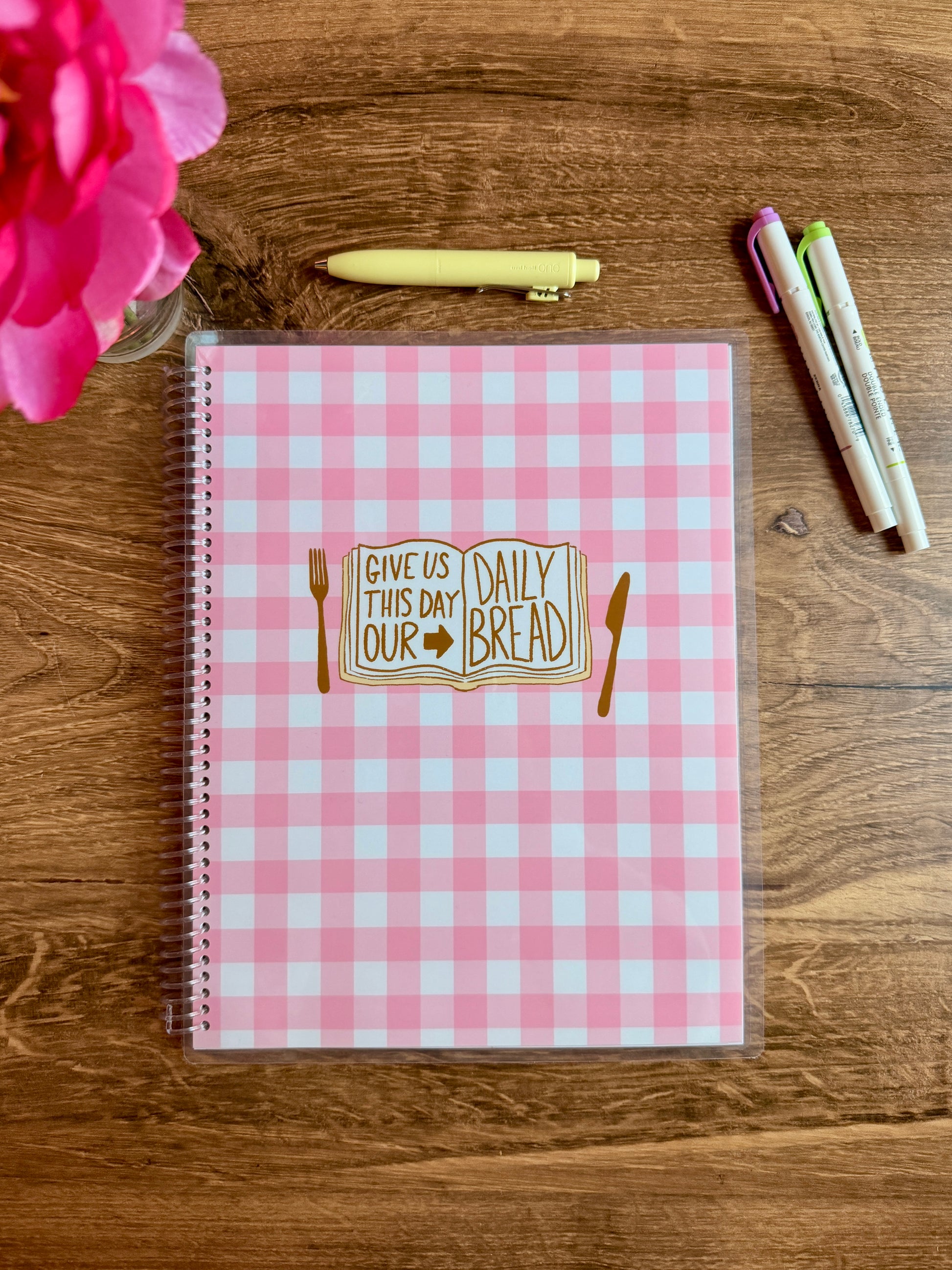 Pink checkered notebook with illustration of a bible that says "give us this day our daily bread" with fork and knife on a wooden surface with a flower and pens.