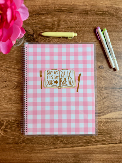 Pink checkered notebook with illustration of a bible that says "give us this day our daily bread" with fork and knife on a wooden surface with a flower and pens.