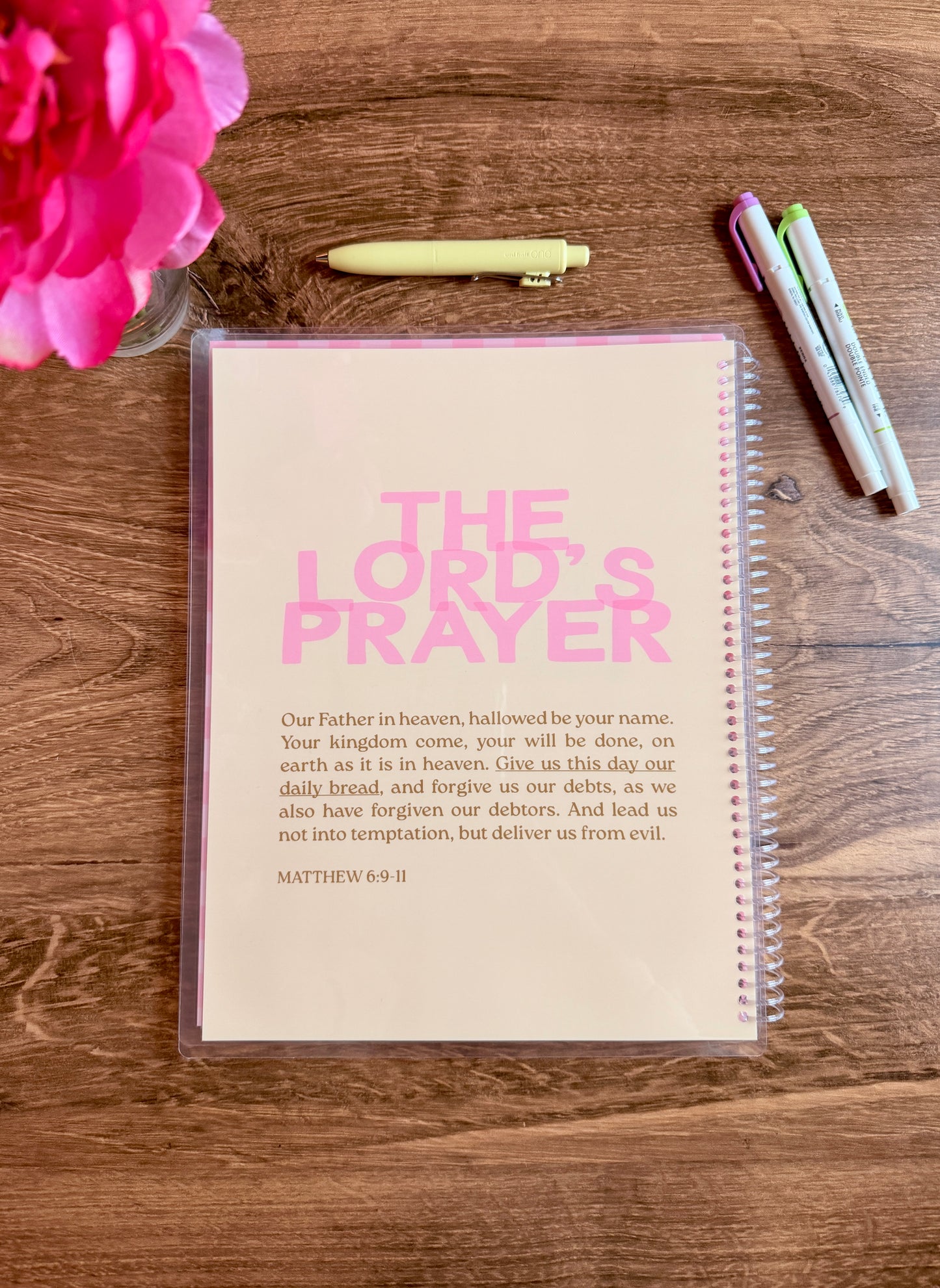 Notebook with 'The Lord's Prayer' printed on it, placed on a wooden surface with pens and a pink flower.