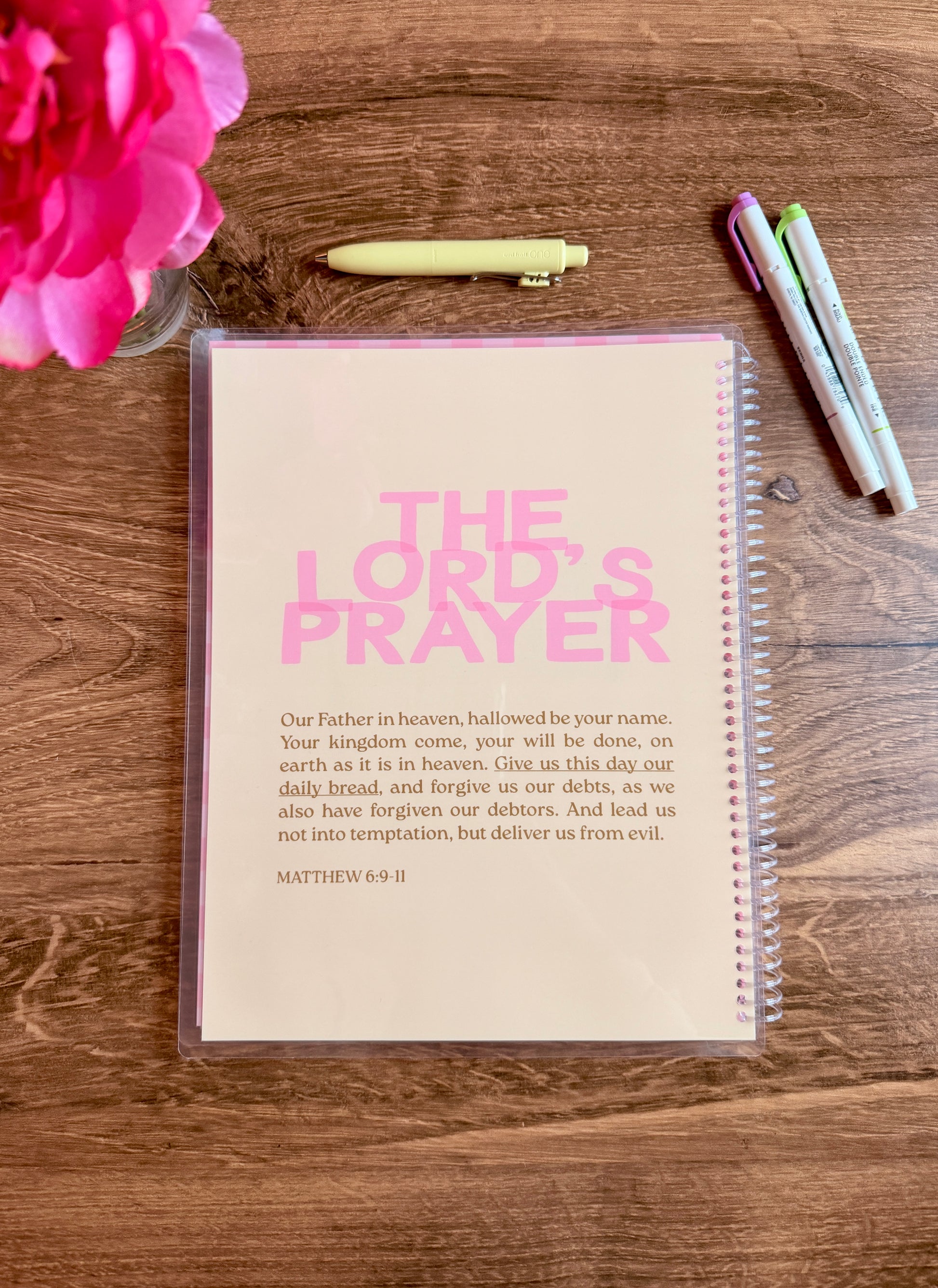 Notebook with 'The Lord's Prayer' printed on it, placed on a wooden surface with pens and a pink flower.
