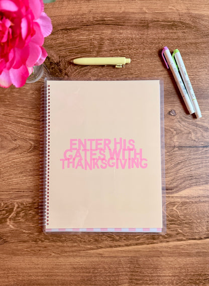 Notebook with 'Enter His Gates with Thanksgiving' text on a wooden surface with a pink flower and markers.