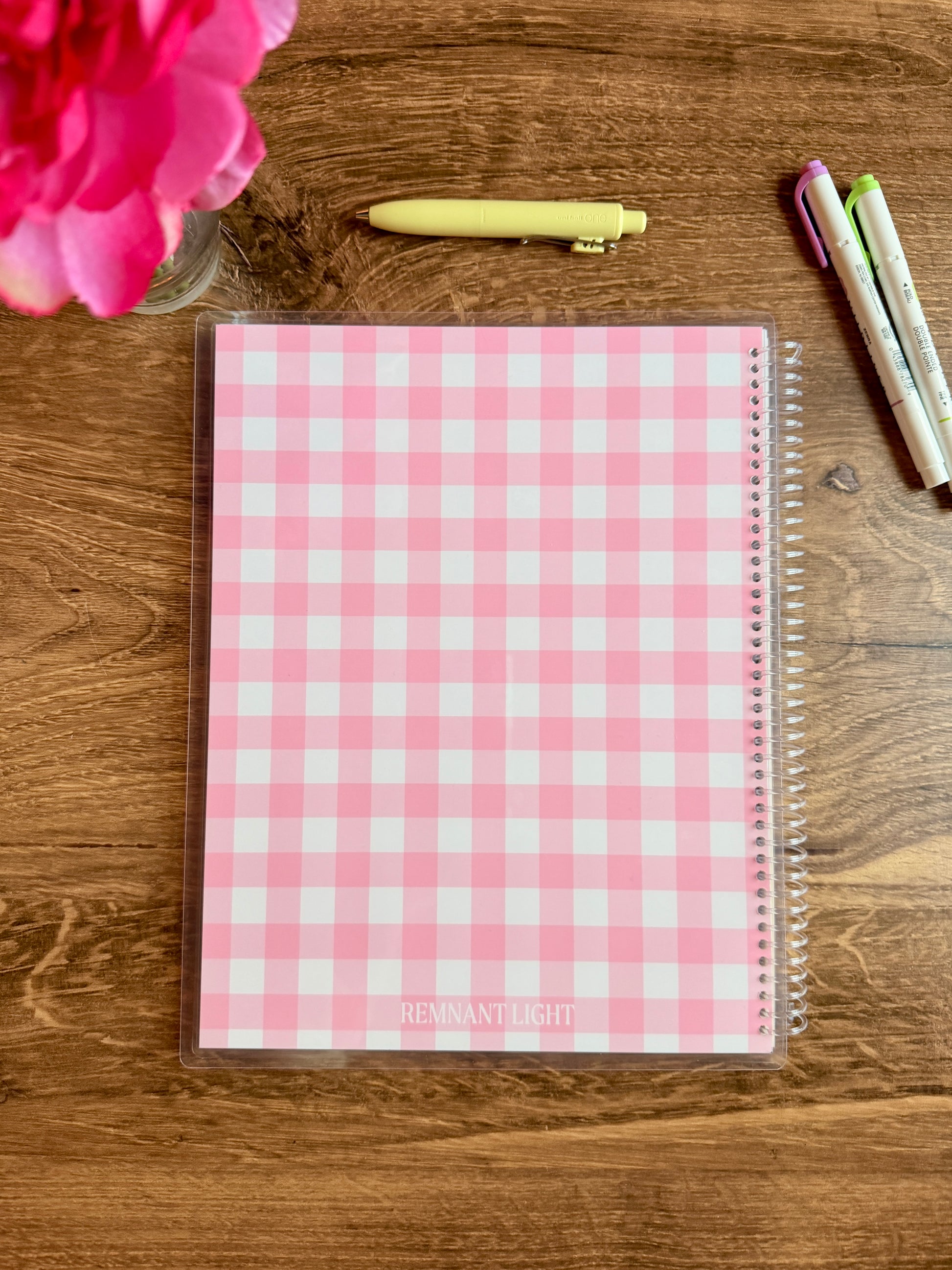 Pink checkered notebook on a wooden surface with a pen and flowers.