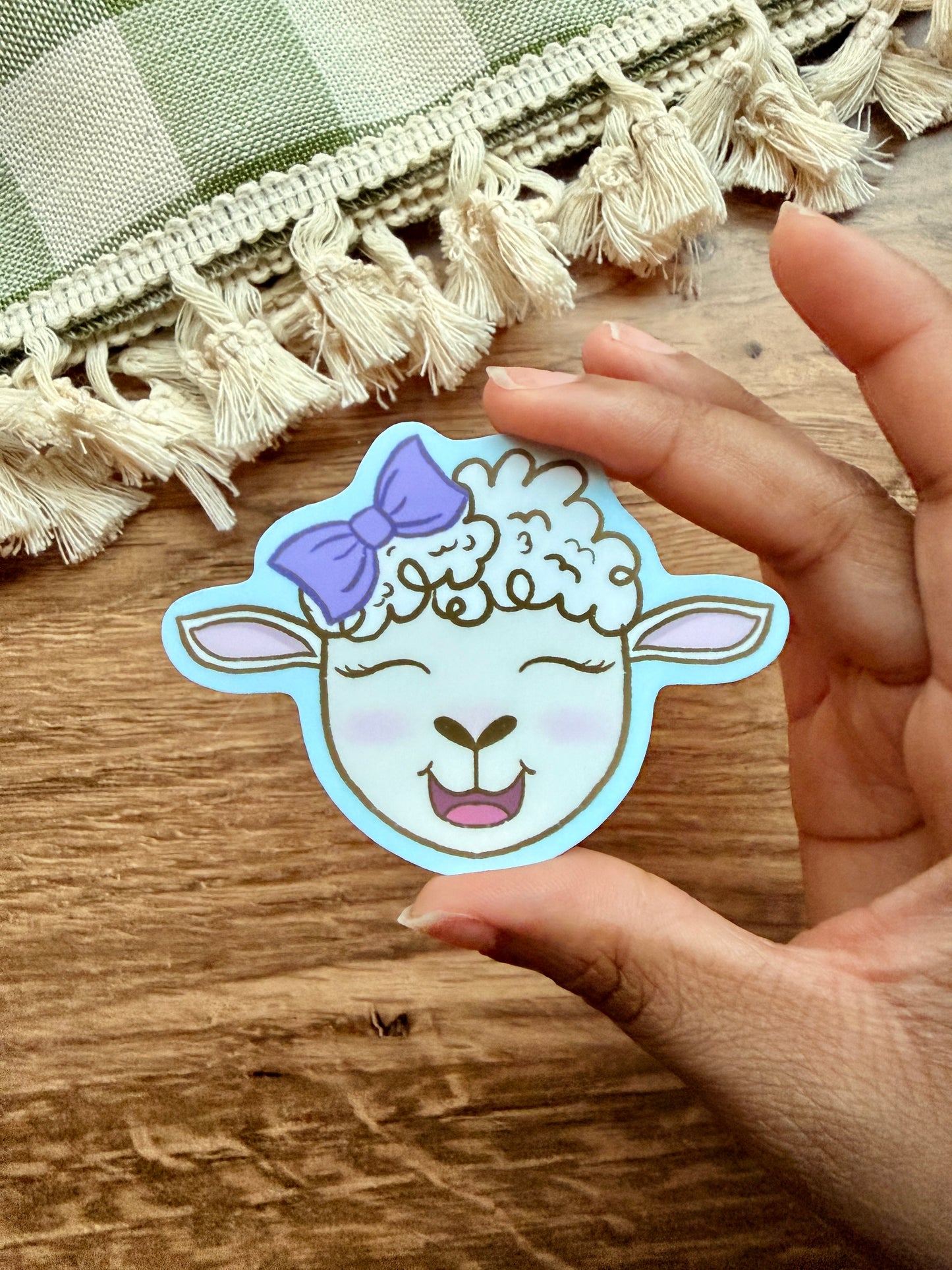 Joyful Little Lamb sticker featuring a cute sheep with a purple bow, held in a hand against a wooden surface.