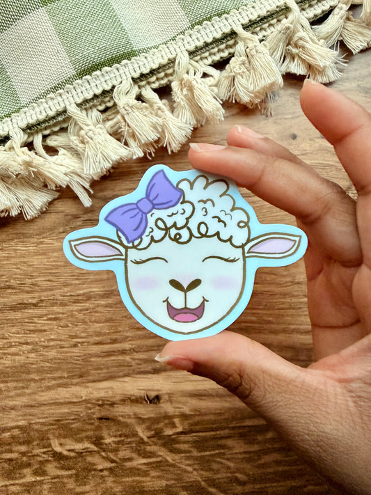 Joyful Little Lamb sticker featuring a cute sheep with a purple bow, held in a hand against a wooden surface.